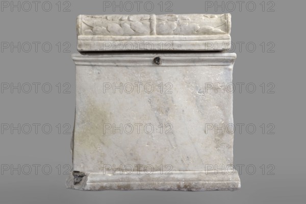 Urn with a depiction of swans, unknown Roman workshop. Main plate: the urn's box is trapezoidal, tapering towards the top and placed on a profiled base. The front surface is sparsely decorated; the main area is occupied by a tabula ansata whose ears are decorated with palmettes. Below the inscription field are two swans in deep relief, facing each other with strongly arched necks, appearing to take flight with outstretched wings touching the inscription frame. The upper part is crowned by a profiled cornice; on the inner upper edge a rectangular protrusion for securing the lid is visible. Side walls: no decoration; metal pins visible under the cornice on both sides. Lid: original missing. Dated to the 2nd half of the 1st century AD probably 3rd quarter of the 1st century AD, Roman Empire 27 BC–476, findplace: Rome. Urn Roman. Weight 45.5 kg., A rectangular stone structure with a flat top and carved upper edge sits against a plain gray background; the stone surface is mostly white with ...