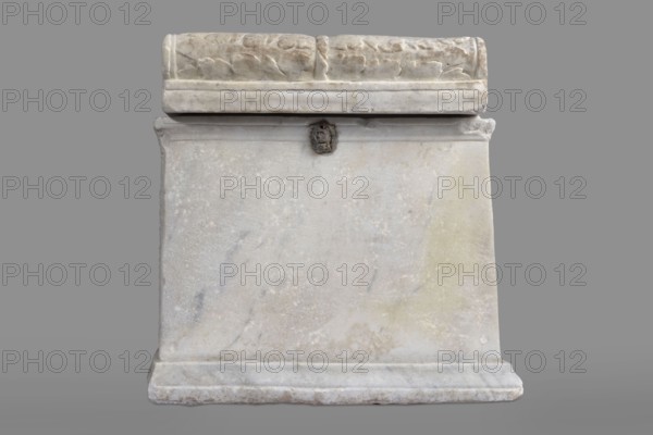Urn with a depiction of swans, unknown Roman workshop. Sepulchral funerary architecture. Main plate: trapezoidal urn tapering toward the top, placed on a profiled base. Front decoration sparse; main surface occupied by a tabula ansata with palmette-decorated ears. Below the inscription field are two swans in deep relief, facing each other with strongly arched necks and outstretched wings touching the inscription frame. Upper part crowned by a profiled cornice; rectangular protrusion on the inner upper edge used to secure the lid. Side walls: no decoration. Metal pins visible under the cornice on both sides. Lid: original missing, currently fitted with a different lid. Dated to the 2nd half of the 1st century AD probably 3rd quarter of the 1st century AD — Roman Empire 27 BC–476. Place of origin/find: Rome. Urn Roman. Weight: 45.5 kg., A rectangular stone structure with a carved lid rests on a flat base, showing a weathered pale surface in shades of white, light gray, cream, and hints o...