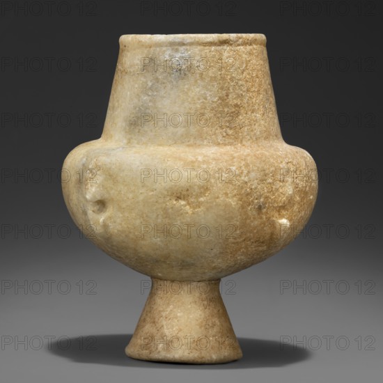 Stone vessel – kandila, Body in the shape of a flattened hemisphere with four evenly spaced, ribbed, vertical ribs. The ribs have round holes suspended through their centers and slightly protrude into the surface of the body. Narrow, flattened shoulders. Neck clearly defined, wide, and conical. Spout in the form of a delicately defined roller running around the rim of the vessel. Interior hollowed out in the neck area with a shallow, semicircular recess protruding into the body; the remaining part of the belly is not hollowed out. 3200–2800 BC Early Cycladic I period, Grotta-Pelos culture, Cyclades. Vessel. Height 11.5 cm, width 9 cm, weight 0.734 kg., An off-white to beige stone vessel with a rounded body that tapers to a short flared pedestal base, a narrower cylindrical neck rising from the shoulder, a matte slightly rough surface with visible grain and subtle mottling in creamy, tan, and light brown tones, a few shallow depressions on the body, and a soft shadow cast beneath it on ...