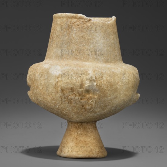 Stone vessel kandila, Body shaped as a flattened hemisphere with four evenly spaced, ribbed vertical ribs. The ribs have round holes through their centers and slightly protrude into the body surface. Narrow, flattened shoulders; clearly defined, wide conical neck. Spout formed by a delicately defined roller around the rim. Interior hollowed in the neck area with a shallow semicircular recess protruding into the body; remaining part of the belly is solid not hollowed. Early Cycladic I period, Grotta–Pelos culture, 3200–2800 BC, Cyclades. Vessel; height 11.5 cm, width 9 cm, weight 0.734 kg., A pale beige stone vessel with a bulbous body, a narrower cylindrical neck with an uneven rim, and a flared conical foot, showing a mottled surface texture and subtle variations of beige, cream, and light brown, set against a dark gray background with a soft shadow beneath.