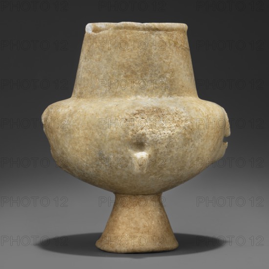 Stone vessel – kandila, Body in the shape of a flattened hemisphere with four evenly spaced, ribbed, vertical handles. The handles have round holes pierced through their centers and slightly protrude into the surface of the body. Narrow, flattened shoulders. Neck clearly defined, wide, and conical. Spout formed by a delicately defined roll running around the rim of the vessel. Interior hollowed out in the neck area with a shallow, semicircular recess protruding into the body; the remaining part of the belly is not hollowed out. 3200–2800 BC Early Cycladic I period, Grotta–Pelos culture, Cyclades. Vessel. Height 11.5 cm, width 9 cm, weight 0.734 kg., A single stone vessel sits upright against a dark gray background, with a rounded body on a flared pedestal base and a short cylindrical neck with a slightly uneven rim; the surface shows a textured, weathered appearance with rough patches and small chips, and the visible colors are beige, light tan, off-white, and hints of pale brown, cast...