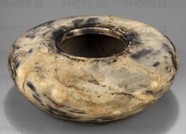 Stone vessel, Stone vessel in the shape of a bird's nest with flat, broad shoulders, a small spout opening not bordered by any protrusion diameter 6.1 cm, walls narrowing in an arch towards a flat, undivided foot. Ca. 2890–2613 BC; Early Dynastic Period, 2nd–3rd Dynasty, Egypt. Vessel; height 6.5 cm, width 15.3 cm, weight 1.0 kg. Repossession provenance probably. Egyptian art; stone vessel; vessels with a separate spout no spout., A rounded, shallow vessel with a wide central opening sits on a neutral surface; its smooth surface shows a mottled pattern of creamy beige, off-white, gray, black, and touches of brown with areas of darker smudging and subtle lighter veins, and a small spot of muted red is visible near the lower edge.