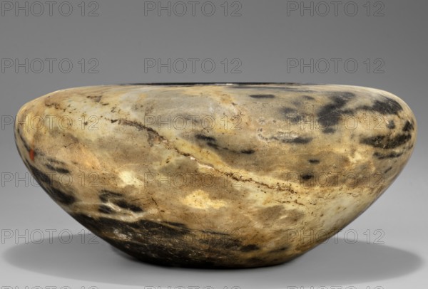 Stone vessel, Stone vessel in the shape of a bird's nest with flat, broad shoulders and a small unbordered spout opening diameter 6.1 cm. Walls narrow in an arch toward a flat, undivided foot. Ca. 2890–2613 BC; Early Dynastic Period, 2nd–3rd Dynasty, Egypt. Vessel; height 6.5 cm, width 15.3 cm, weight 1.0 kg., A smooth, rounded stone bowl with an oval opening resting on a flat surface, displaying a mottled surface of beige, tan, brown, black, gray, and hints of cream; the bowl shows irregular darker patches and streaks across its exterior, subtle veins and textured markings, and a soft shadow beneath it on a neutral gray background.