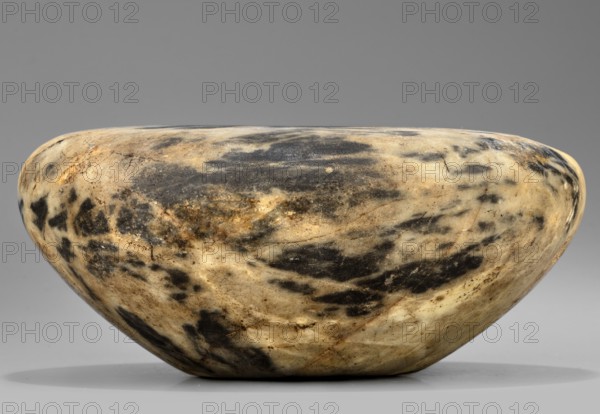 Stone vessel, stone, in the shape of a bird's nest with flat broad shoulders and a small unbordered spout opening diameter 6.1 mm, walls narrowing in an arch toward a flat undivided foot; ca. 2890–2613 BC, Early Dynastic Period, 2nd–3rd Dynasty, Egypt; vessel; height 6.5 cm, width 15.3 cm, weight 1.0 kg., A smooth, rounded stone bowl with a low, wide profile sits against a plain background; its surface shows a marbled pattern of creamy beige and light tan mixed with patches and streaks of dark gray and black, with subtle areas of brownish discoloration and faint hairline cracks, and the lighting creates soft highlights and gentle shadows along its curved contours.