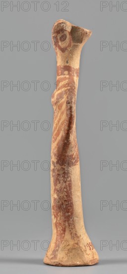 Female idol, unknown Mycenaean workshop. Votive female figurine in the shape of the letter phi—legs form a cylindrical base widening downward skirt?, arms and torso enclosed in a circle, breasts schematically marked, long cylindrical neck, flat head. Folds of garments, facial features, and eyes simplified using brown paint or varnish; painted surface worn in places. Circa 1400–1200 BC previously dated 1600–1100 BC. Provenance: Mycenae, Greece. Figurine votive offering. Height 12.7 cm; width 4.6 cm; depth 2.8 cm; weight 0.05 kg., A tall, narrow ceramic figurine photographed against a plain gray background, showing an elongated form with a flared base and a tapered top. The surface displays a mix of earthy beige and reddish-brown tones, with the reddish-brown appearing as painted bands and diagonal stripes along the body and head area. The figure has a rounded head with a suggestion of circular painted elements at the top, a slight protrusion at the chest, and a gently curving silhouette...