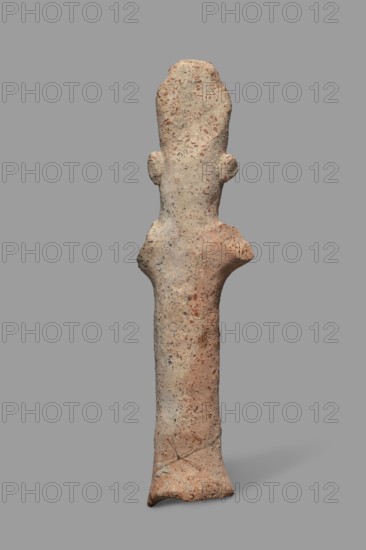Votive figurine, Cyprus; male figurine with tiara triple crown, votive offering. Figurine of a man in a high headdress, with a cylindrical body and cylindrical arms raised upwards and outstretched to the sides. The face, neck, and torso are of equal thickness; only a large nose is visible. Large ears are attached to the sides of the head. Dated 750–600 BC formerly 7th–6th century BC. Terracotta figurine, height 14.2 cm, width 5.0 cm., A tall, slender, upright textured object resembling a rough sculpture with an elongated body and a slightly wider top that has two small protrusions on its sides, standing against a plain gray background; the surface shows a mix of beige, light brown, reddish-brown, and small flecks of darker gray, with uneven, pitted texture and some smoother worn areas.