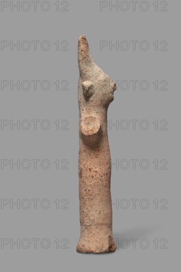 Votive figurine, Cyprus, male figurine wearing a high headdress tiara/triple crown, votive offering. Figurine of a man with a cylindrical body and cylindrical arms raised upwards and outstretched to the sides. The face, neck, and torso are of equal thickness; only a large nose is visible and large ears are attached to the sides of the head. Dated 750–600 BC formerly 7th–6th century BC. Terracotta figurine, height 14.2 cm, width 5.0 cm., A narrow upright elongated sculpture in shades of tan and light brown with speckled darker brown and reddish flecks, standing against a plain gray background; the form tapers to a pointed top with a roughly textured surface, a rounded protrusion on one side near the upper section, a small ear-like bulge lower on the head area, and a circular raised area on the torso; the base is wider and slightly irregular with visible roughness and tiny pits, and the overall piece has a grainy, weathered appearance with subtle variations of beige, tan, brown, and hint...