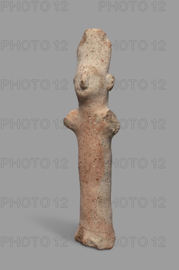Votive figurine, Cyprus, male figurine wearing a tiara triple crown, votive offering. Figurine of a man in a high headdress with a cylindrical body and cylindrical arms raised upwards and outstretched to the sides. The face, neck, and torso are of equal thickness; only a large nose is visible and large ears are attached to the sides of the head. Dated 750–600 BC previously 7th–6th century BC. Terracotta figurine, height 14.2 cm, width 5.0 cm., A weathered standing figurine made of rough textured material with a tall elongated head and simplified facial features, including a protruding nose and small ear shapes, a narrow cylindrical body with short stubby arm-like projections at the shoulders, and an irregular base; the surface shows a mix of pale beige, light tan, and muted reddish-brown tones with speckled darker flecks and some areas of lighter gray, all set against a flat gray background.