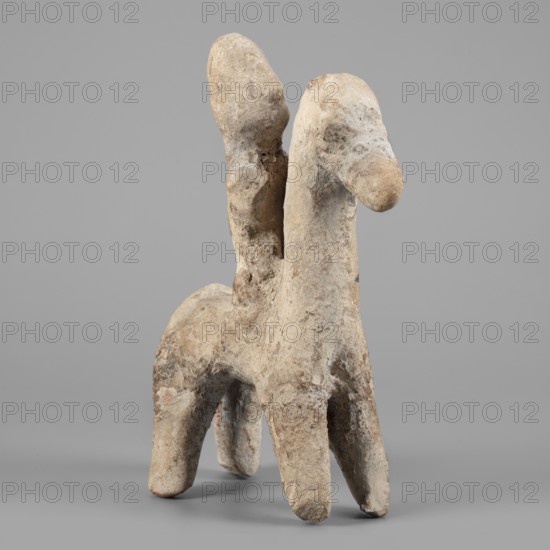 Figurine of a rider on a horse. The entire piece is schematic, the overall shape is highlighted, lacking any details. The horse is small, with a short torso, cylindrical, slightly tapering legs, and an unmarked tail. The neck is very long and vertical, the muzzle is large, the eyes are slightly convex, the ears are small, and the mane is short and trimmed. The rider sits upright, close to the horse's neck. The torso is short, the legs and arms are reflected. The head is large, with a clearly defined profile; the nose is particularly prominent, the eyes are unmarked, and at the back of the head is a vertically sloping, adjacent ridge that can be interpreted as a helmet plume. The helmet itself is not marked, only the overdeveloped top of the head suggests its presence. Dated 750–600 BC previously 7th–6th century BC. Origin: Cyprus. Type: figurine. Dimensions: height 14.9 cm, width 11.1 cm, depth 4 cm., A rough, weathered sculptural form resembling a two-headed quadruped stands on a smoo...