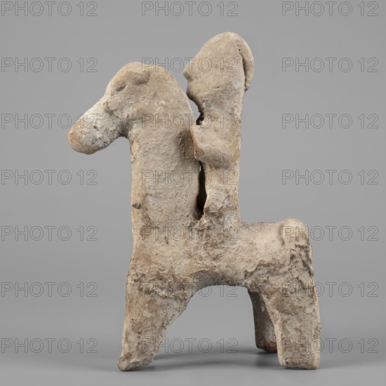 Figurine of a rider on a horse, Cyprus. The entire piece is schematic, the overall shape is emphasized and lacks detail. The horse is small with a short torso, cylindrical slightly tapering legs, and an unmarked tail. The neck is very long and vertical; the muzzle is large; the eyes are slightly convex; the ears are small; the mane is short and trimmed. The rider sits upright, close to the horse's neck; the torso is short; the legs and arms are indicated. The head is large with a clearly defined profile and a particularly prominent nose; the eyes are unmarked. At the back of the head is a vertically sloping adjacent ridge interpreted as a helmet plume the helmet itself is not marked, only the overdeveloped top of the head suggests its presence. Date: 750–600 BC previously dated 7th–6th century BC. Type: figurine. Dimensions: height 14.9 cm, width 11.1 cm, depth 4 cm. Creator: unknown., A roughly textured sculptural object with an off-white to beige surface and patches of light brown, s...