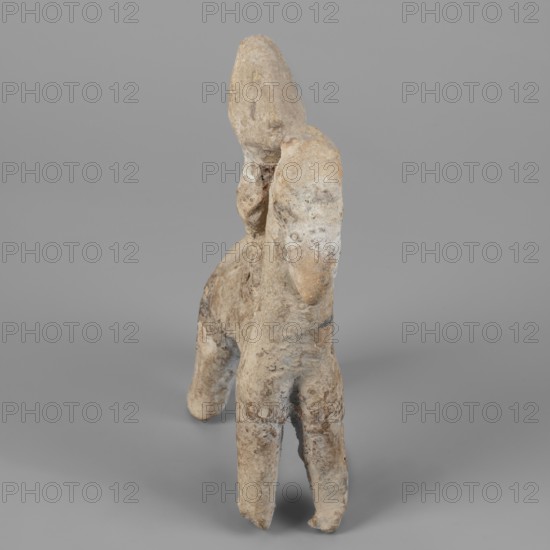 Figurine of a rider on a horse, Cyprus. The entire piece is schematic, the overall shape is emphasized and lacks fine detail. The horse is small with a short torso, cylindrical slightly tapering legs, and an unmarked tail. The neck is very long and vertical; the muzzle is large; the eyes are slightly convex; the ears are small; the mane is short and trimmed. The rider sits upright, close to the horse’s neck; the torso is short; the legs and arms are indicated. The head is large with a clearly defined profile and a particularly prominent nose; the eyes are unmarked. A vertically sloping adjacent ridge at the back of the head may indicate a helmet plume, though the helmet itself is not marked, only an overdeveloped top of the head suggests its presence. Dated 750–600 BC previously dated 7th–6th century BC. Figurine; height 14.9 cm, width 11.1 cm, depth 4 cm., A small weathered sculpture of a figure mounted on an animal, shown against a plain gray background, with the sculpture displaying...