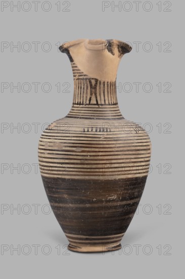 Oinochoe, Geometric oinochoe with a low foot, a gradually widening belly, distinct shoulders, a slender neck, and a tripartite rim characteristic of this vessel form. The vase runs from the shoulder to the rim. Halfway up the belly it is covered with black finish; the rest is decorated with closely spaced parallel horizontal lines encircling the vessel. The central part of the neck is decorated with vertical stripes. Oinochoe used for drawing and pouring wine Greek: oinos - wine and is a typical Greek vessel. Late 8th century BC sometimes dated to the end of the 8th–beginning of the 7th century BC; described as an Italian imitation of Proto-Corinthian, Cumae workshop, Southern Italy Italy. Vessel oinochoe. Weight 0.476 kg; height 23.2 cm; diameter 12.2 cm., A ceramic vessel with a flared rim and narrow neck leading to a rounded body, displayed against a plain gray background; the vessel shows alternating horizontal bands in black and light beige, with a darker black banding toward the ...