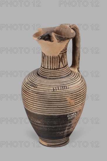 Oinochoe, Geometric, end of the 8th century BC possibly end of 8th–beginning of 7th century BC; described as an Italian imitation of a Proto-Corinthian/Cumae workshop. Geometric oinochoe with a low foot, a gradually widening belly, distinct shoulders, a slender neck, and a tripartite rim. The vase runs from the shoulder to the rim. Halfway up the belly it is covered with black finish; the rest is decorated with closely set parallel horizontal lines encircling the vessel. The central part of the neck is decorated with vertical stripes. Function: vessel for drawing and pouring wine Greek: oinos. Southern Italy Cumae. Vessel type: oinochoe. Weight 0.476 kg; height 23.2 cm; diameter 12.2 cm., A ceramic jug with a rounded body, narrow neck, flared rim, and a single curved handle is shown against a neutral background; the jug has a beige base color with black horizontal bands encircling the body and neck, black painted accents near the rim and handle, and areas of darker brown and worn beige...