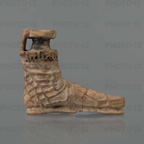 Foot-shaped aryballos, Ionian workshop; aryballos, antique vessel; yellow-orange clay with mica; matte black engobe on the surface. Aryballos in the shape of a foot shod in a sandal with a thick sole connected to braided leather straps. The foot ends above the ankle and merges into a narrow neck terminating in an outward-turned rim, decorated from above with radiating lines. A wide, ribbon-like handle is attached vertically to the spout, on which there is an image of a rooster surrounded by dots. Preservation condition: the front part of the foot is filled with plaster. Dated end of the 6th–beginning of the 5th century BC. Ionia, Turkey/Greece., A sculpted object in the shape of a boot shown in profile on a neutral gray background, with a handle-like extension at the top near the ankle; the surface of the boot is textured with a pattern resembling scales or stitched segments and has an uneven, weathered appearance with visible cracks and roughness; the colors visible are tan, beige, br...