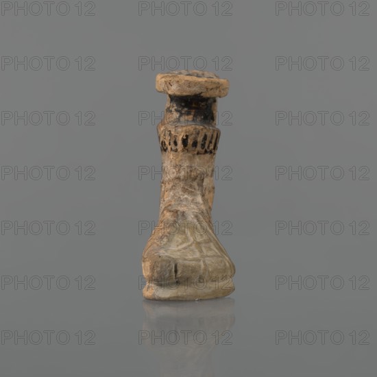 Foot-shaped aryballos, Ionian workshop, aryballos, antique vessel. Yellow-orange clay with mica; matte black engobe on the surface. Shaped as a foot shod in a sandal with a thick sole connected to braided leather straps. The foot ends above the ankle and merges into a narrow neck terminating in an outward-turned rim, decorated from above with radiating lines. A wide, ribbon-like handle is attached vertically to the spout, bearing an image of a rooster surrounded by dots. Preservation: the front part of the foot is filled with plaster. Date: end of the 6th – beginning of the 5th century BC. Findspot: Ionia, Turkey/Greece., A small carved object resembling a stylized foot or shoe stands upright against a plain gray background, showing a weathered surface with a mix of beige, tan, brown, and darker blackened areas; the sole and toe area are rounded and display worn texture and subtle carved lines, the midsection tapers upward with irregular rough patches and lighter beige highlights, and ...