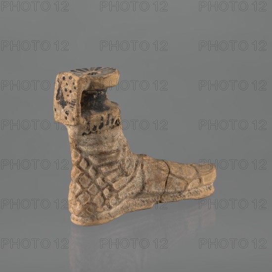 Foot-shaped aryballos, aryballos, antique vessel. Yellow-orange clay with mica; matte black engobe on the surface. Shaped as a foot shod in a sandal with a thick sole connected to braided leather straps. The foot ends above the ankle and merges into a narrow neck terminating in an outward-turned rim, decorated from above with radiating lines. A wide, ribbon-like handle is attached vertically to the spout, bearing an image of a rooster surrounded by dots. Preservation condition: the front part of the foot is filled with plaster. End of the 6th–beginning of the 5th century BC. Ionia, Turkey/Greece., Ionian workshop., A worn sculpted object resembling a boot shape sits against a plain gray background, showing a textured surface with carved patterns and a few incised lines. The piece displays a mottled palette of beige, tan, and light brown with darker brown and black smudges and spots in several areas, especially near the upper section. The surface appears rough and weathered with visible...