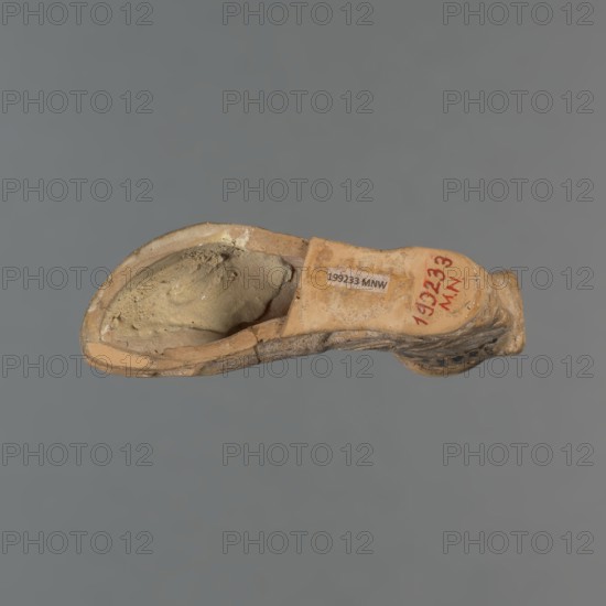 Foot-shaped aryballos, aryballos, antique vessel. Yellow-orange clay with mica; matte black engobe on the surface. Shaped like a foot shod in a sandal with a thick sole connected to braided leather straps. The foot ends above the ankle and merges into a narrow neck terminating in an outward-turned rim, decorated from above with radiating lines. A wide, ribbon-like handle is attached vertically to the spout, on which there is an image of a rooster surrounded by dots. Preservation condition: the front part of the foot is filled with plaster. End of the 6th–beginning of the 5th century BC. Made in an Ionian workshop; found in Ionia/Turkey/Greece., A worn, hollow object shaped like a curved vessel rests against a plain gray background; the object shows a beige and tan exterior with patches of darker brown and lighter cream tones, a rough textured inner cavity filled with a sandy, muted yellow-beige material, and areas of surface abrasion and small chips along the rim and edges.