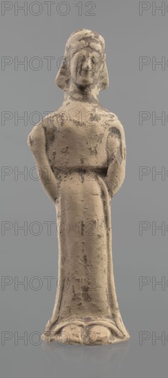 Terracotta figurine of a woman, unknown Boeotian workshop. Mold-pressed terracotta figurine of a standing woman in a long chiton. Head set on a thick neck; oval face with a large nose and almond-shaped eyes. Arms bent at the elbows, forearms raised so that the hands are pressed against the shoulders. Right hand holds a cylindrical object probably a fragment of a longer element. Traces of brown and red polychrome. Condition: left hand broken off, surface worn. Ca. 550 BC. Boeotia. Figurine, height 23 cm, width 7 cm., A standing figurine with a worn, textured surface in beige and light tan with darker grayish-brown patches, showing a simplified head with faint facial features and stylized hair, narrow shoulders and arms held close to the body, a long straight torso and skirt that flares slightly at the base, and a smooth neutral gray background.