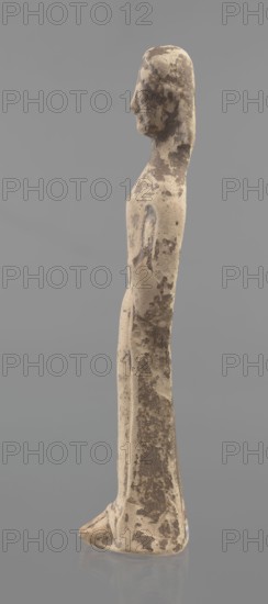 Terracotta figurine of a woman, unknown Boeotian workshop. Female mold-pressed figurine. Standing woman in a long chiton. Head set on a thick neck. Oval face with a large nose and almond-shaped eyes. Arms bent at the elbows, forearms raised so that the hands are pressed against the shoulders. In the right hand is a cylindrical object probably a fragment of a longer element. Traces of brown and red polychrome. Condition: left hand broken off, surface worn. Circa 550 BC, Boeotia. Figurine. Height 23 cm, width 7 cm., A slender standing figure rendered in a weathered beige material with darker brown patches across its surface, shown in profile against a solid gray background; the figure has an elongated head and neck, a simplified facial profile, narrow shoulders, an arm indicated along the side, and a long straight body that tapers to a flat base with visible textured wear and mottled discoloration in beige and brown.
