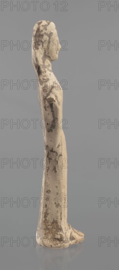 Terracotta figurine of a woman, unknown Boeotian workshop. Female mold-pressed figurine standing in a long chiton. Head set on a thick neck; oval face with a large nose and almond-shaped eyes. Arms bent at the elbows, forearms raised with hands pressed against the shoulders. Right hand holds a cylindrical object probably a fragment of a longer element. Traces of brown and red polychrome. Condition: left hand broken off, surface worn. Ca. 550 BC. Boeotia. Figurine; height 23 cm, width 7 cm., A slender standing figure viewed in profile, rendered in a weathered beige material with irregular darker brown patches and spotting across the surface; the figure has an elongated head with a pronounced nose, a long neck, and narrow shoulders leading into a straight, columnar torso and skirted lower body, with the suggestion of feet at the base; the surface shows rough texture, chips, and worn areas, and the background is a smooth mid-gray.
