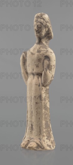 Terracotta figurine of a woman, unknown Boeotian workshop. Female mold-pressed terracotta figurine. Standing woman in a long chiton. Head set on a thick neck. Oval face with a large nose and almond-shaped eyes. Arms bent at the elbows, forearms raised so that the hands are pressed against the shoulders. In the right hand is a cylindrical object probably a fragment of a longer element. Traces of brown and red polychrome. Condition: left hand broken off, surface worn. Ca. 550 BC, Boeotia. Figurine; height 23 cm, width 7 cm., A worn, upright figurine with a rounded head and simplified facial features, standing on a flared base; the figure wears a long, straight garment with subtle folds and has arms bent at the elbows with hands near the torso. The surface shows irregular patches and speckles of darker discoloration against a lighter, beige-toned material, with visible rough texture and small pitting. The overall colors visible are beige and darker brownish-gray, set against a neutral gra...