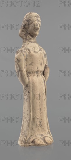 Terracotta figurine of a woman, unknown Boeotian workshop. Mold-pressed terracotta figurine. Standing woman in a long chiton. Head set on a thick neck; oval face with a large nose and almond-shaped eyes. Arms bent at the elbows, forearms raised with the hands pressed against the shoulders. Right hand holds a cylindrical object probably a fragment of a longer element. Traces of brown and red polychrome. Condition: left hand broken off, surface worn. ca. 550 BC, Boeotia. Height 23 cm, width 7 cm., A standing figurine made of a light beige material with a textured, slightly rough surface and patches of darker brown discoloration, showing a simplified human form with a rounded head, indistinct facial features, shoulder-length hair suggestion, slender neck, narrow shoulders, and long arms positioned at the sides with hands resting near the hips; the body is clothed in a long, straight garment that falls to a flared base with gentle folds and rounded contours, and the entire figure is shown ...