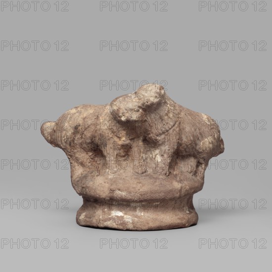 Two dogs, Two dogs shown schematically on a round, profiled base. The dogs are shown standing, with their heads touching. 332–30 BC; Ptolemaic Period, Tell Atrib; figurine; height 5.3 cm, width 7.1 cm., A small weathered clay sculpture depicting a group of animal-like forms atop a rounded base, with rough textured surfaces and visible wear; colors include shades of brown, tan, beige, and hints of off-white, set against a neutral gray background.