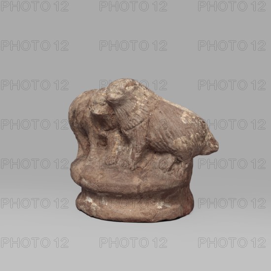 Two dogs, Two dogs shown schematically on a round, profiled base. The dogs are shown standing, with their heads touching. Ptolemaic Period, 332–30 BC; Tell Atrib. Figurine; height 5.3 cm, width 7.1 cm., A small weathered sculpture of two animals standing close together on a rounded base, both showing textured fur and rounded heads; the piece appears rough and eroded with uneven surfaces and small patches of lighter wear. The visible colors are brown, tan, beige, and hints of off-white against a neutral gray background.