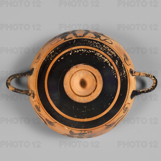 Kylix with a depiction of a mermaid and sphinxes, Griffin-Bird Painter, fantasy creatures, sphinxes, sirens; Greek pottery, 570–550 BC previously dated ca. 545 BC, Attica; vessel kylix; weight 0.314 kg, A round ceramic vessel seen from above with a central recessed area, a wide black circular band with scattered small chips revealing the underlying color, concentric rings, and an outer orange surface decorated with black painted motifs; the vessel has two loop handles on opposite sides showing wear, and visible colors include orange, black, and small areas of lighter tan where the surface is worn.