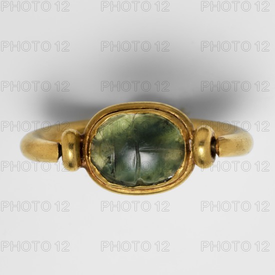 Ring with a scarab and a cryptogram of Amun;, Materials/technique: gold, jewelry; type: scarab amulet ring. Decorations: plant motifs; underside of the base is engraved — a bull in right profile with an ankh sign above it; a tuft of vegetation in front of the bull on the right; at the bottom a double horizontal line interrupted by three lines mat, probably a cryptogram of the name Amun. Iconography: Amun mythology, ankh. Date/place: c. 1550–c. 1295 BC, New Kingdom, 18th Dynasty, Egypt., A close-up of a gold ring with a rounded band and a bezel-set oval green gemstone; the gold has a warm yellow tone with slightly worn surfaces and small rounded gold accents on either side of the stone, and the gemstone shows varying shades of green with internal inclusions and a glossy, slightly domed surface against a neutral light gray background.