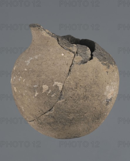 Vessel with a rounded bottom, unknown northern Mesopotamian workshop, vice-free vessel, Early Jazira III period ca. 2600–ca. 2350 BC / Early Dynastic III period ca. 2600–ca. 2350 BC. Thick-walled vessel with a rounded bottom and a bulging belly with short, arched shoulders merging into a low neck terminating in a slightly inclined straight spout. ca. 2600–ca. 2350 BC; Tell Abu Hafur northeastern Syria, Northern Mesopotamia Khabur basin. Vessel; height 15 cm, width 13.2 cm, rim diameter 6.9 cm., A round, weathered clay vessel with a narrow, slightly upturned spout and a large irregular break at the rim, showing rough, uneven interior edges; the surface is textured with pitting, small chips, and areas of flaking. The object displays a palette of muted beige and tan tones across most of its body, with patches of darker gray and black near the broken rim and scattered lighter off-white speckles on the exterior. The background is a uniform medium gray.