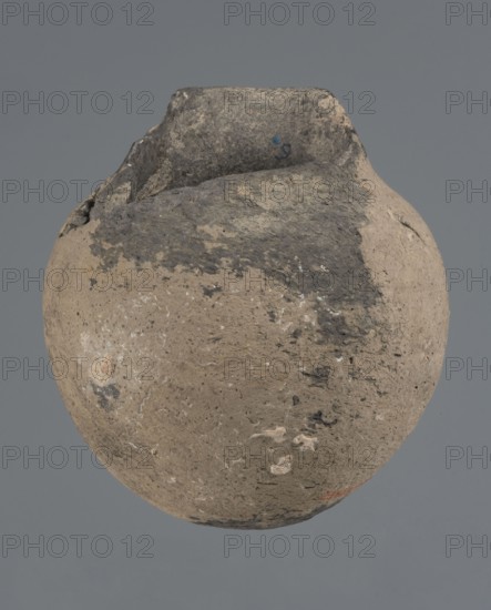 Vessel with a rounded bottom, unknown northern Mesopotamian workshop, vice-free vessel, Early Jazira III period ca. 2600–ca. 2350 BC / Early Dynastic III period ca. 2600–ca. 2350 BC. Thick-walled vessel with a rounded bottom and a bulging belly with short, arched shoulders merging into a low neck terminating in a slightly inclined straight spout. Ca. 2600–ca. 2350 BC; Tell Abu Hafur northeastern Syria, Northern Mesopotamia Khabur basin. Vessel; height 15 cm, width 13.2 cm, rim diameter 6.9 cm., A rounded, weathered clay vessel with a rough surface and an irregular rim is shown against a plain gray background, displaying areas of beige and light brown clay with patches of darker gray and black discoloration, small whitish spots and mineral-like flecks, and uneven texture with chips and pitted marks.