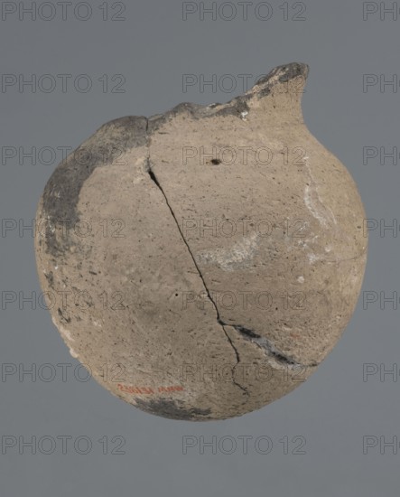 Vessel with a rounded bottom, unknown northern Mesopotamian workshop; vice-free, thick-walled vessel with a rounded bottom and a bulging belly with short, arched shoulders merging into a low neck terminating in a slightly inclined straight spout. Early Jazira III / Early Dynastic III period ca. 2600–ca. 2350 BC. From Tell Abu Hafur, Al-Jazira Khabur basin, northern Mesopotamia. Material: ceramic. Height 15 cm; width 13.2 cm; rim diameter 6.9 cm., A rounded, weathered pottery vessel with a rough, uneven surface in shades of tan, beige, and gray suspended against a plain gray background, showing several visible cracks and a chipped rim with darker gray discoloration, small pits and speckles across the body, and a few areas of lighter residue or mineral deposits.