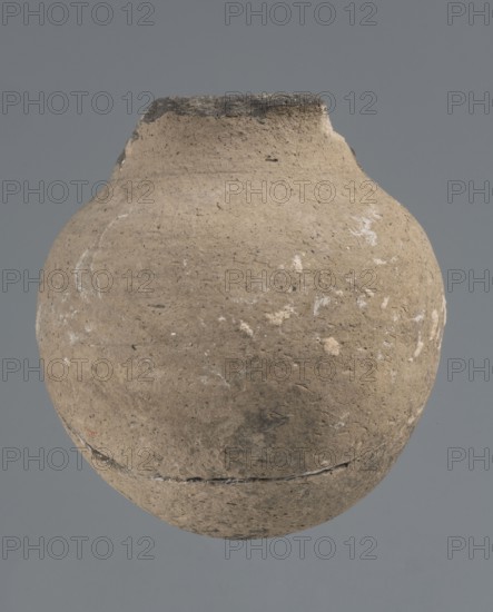 Vessel with a rounded bottom, unknown northern Mesopotamian workshop; vice-free vessels; Early Jazira III period ca. 2600–ca. 2350 BC / Early Dynastic III period ca. 2600–ca. 2350 BC. Thick-walled vessel with a rounded bottom and a bulging belly with short, arched shoulders merging into a low neck terminating in a slightly inclined straight spout. ca. 2600–ca. 2350 BC; Tell Abu Hafur northeastern Syria, Northern Mesopotamia Khabur basin. Vessel; height 15 cm, width 13.2 cm, rim diameter 6.9 cm., A rounded, earthen vessel with a narrow, slightly irregular neck and worn rim sits centered against a plain gray background; the vessel surface is rough and textured with a dominant beige-brown color, darker brown and gray patches, and scattered pale white areas and flecks, with visible surface pitting and a horizontal seam around the lower portion.