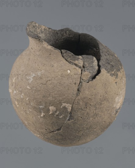 Vessel with a rounded bottom, unknown northern Mesopotamian workshop; vice-free vessels; Early Jazira III / Early Dynastic III period ca. 2600–ca. 2350 BC. Thick-walled vessel with a rounded bottom and a bulging belly with short, arched shoulders merging into a low neck terminating in a slightly inclined straight spout. Tell Abu Hafur northeastern Syria; Northern Mesopotamia Khabur basin. Vessel; height 15 cm, width 13.2 cm, rim diameter 6.9 cm., A roughly spherical clay vessel with an irregular broken rim, showing a cracked and chipped surface; the exterior is mostly light brown with areas of darker brown and some lighter pale spots, and the interior and rim edges appear dark gray to black; the background is a flat medium gray.