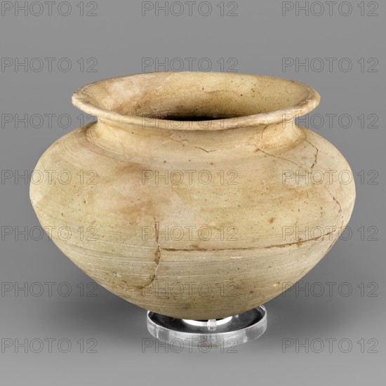 Vessel with an egg-shaped base, unknown Northern Mesopotamian workshop. Vessel for oils and ointments, vice-free. Vessel with an egg-shaped base and a bulging, conically inclined belly, transitioning into short, rounded shoulders with a low, inclined neck terminating in a narrow, outwardly curled rim; on the inside the neck is separated from the rim by a thin engraved line. 1st half of the 3rd millennium BC; Early Jazira III period Early Dynastic III; ca. 2600–ca. 2350 BC. Tell Abu Hafur North-Eastern Syria, Northern Mesopotamia Khabur basin. Vessel. Height 9.2 cm, width 13.3 cm, weight 270 g., A rounded ceramic vessel with a wide flaring rim sits on a clear circular stand against a gray background. The vessel shows a smooth, worn surface with visible hairline cracks and repair lines, and areas of uneven texture. Its colors include pale beige, light tan, warm cream, and small patches of darker brown discoloration. The interior appears darker than the exterior, and the rim has slightly ...