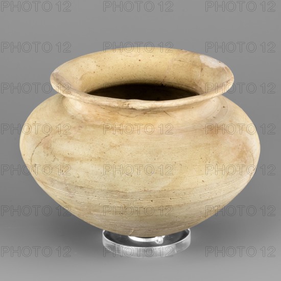 Vessel with an egg-shaped base, unknown Northern Mesopotamian workshop; vessel for oils and ointments, vice-free vessel. Vessel with an egg-shaped base and a bulging, conically inclined belly, transitioning into short, rounded shoulders with a low, inclined neck terminating in a narrow, outwardly curled rim. On the inside, the neck is separated from the rim by a thin engraved line. 1st half of the 3rd millennium BC; Early Jazira III period Early Dynastic III; ca. 2600–ca. 2350 BC. Tell Abu Hafur North-Eastern Syria, Northern Mesopotamia Khabur basin. Vessel; height 9.2 cm, width 13.3 cm, weight 270 g., A rounded, wide-mouthed ceramic vessel sits on a clear circular stand against a plain background; the pot has a bulbous body with a slightly flared rim and visible surface irregularities and faint circular marks from shaping. The visible colors are beige, light tan, cream, pale yellow, and touches of brown, with subtle darker areas inside the opening and slight discolorations and streaks...