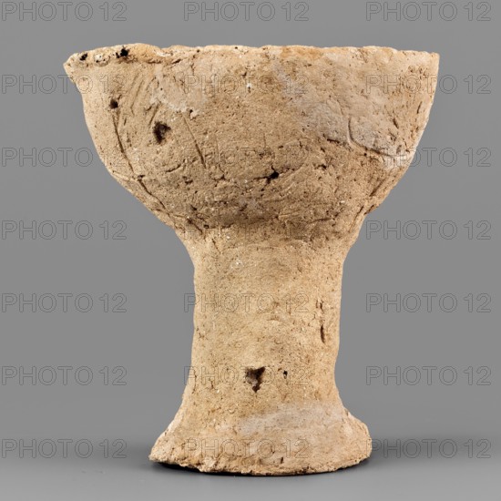 Beaker with engraved decoration, unknown northern Mesopotamian workshop. Hand-made beaker on a thick, cylindrical stem with a poorly defined foot. The bowl is a low form with an irregularly defined rim, glued to the foot. Three schematically engraved representations of human figures are on the bowl. Each torso with head and legs is depicted by a vertical line terminating in a dot at the top and a short arc opening downward at the bottom. The arms, extended upward, are marked by an engraved horizontal line, curved at both ends, which intersects the vertical line of the torso slightly below its midpoint. Anthropomorphic engraved decoration. 1st half of the 3rd millennium BC; Early Jazira III period Early Bronze/Early Dynastic III; ca. 2600–ca. 2350 BC. Tell Abu Hafur northeastern Syria, Khabur basin, Northern Mesopotamia. Vessel, weight 148 g., A roughly shaped, beige-brown clay vessel with a wide, shallow bowl top narrowing into a thick stem and flared base, showing a coarse, grainy sur...