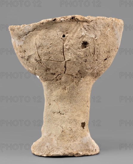 Beaker with engraved decoration, unknown northern Mesopotamian workshop. Hand-made beaker on a thick, cylindrical stem with a poorly defined foot. The bowl is a low form with an irregularly defined rim, glued to the foot. Three schematically engraved human figures on the bowl: the torso with head and legs is a vertical line terminating in a dot at the top and a short arc opening downward at the bottom; the arms, extended upward, are marked by an engraved horizontal line curved at both ends that intersects the torso slightly below its midpoint. 1st half of the 3rd millennium BC; Early Jazira III period Early Bronze Period; Early Dynastic III period; ca. 2600–ca. 2350 BC. Tell Abu Hafur northeastern Syria, Khabur basin; Northern Mesopotamia Khabur basin. Vessel; weight 148 g., A weathered, textured cup-like vessel with a wide bowl atop a narrow stem and a flared base, shown against a plain gray background. The object has an uneven, rough surface with visible cracks, pits, and small holes...