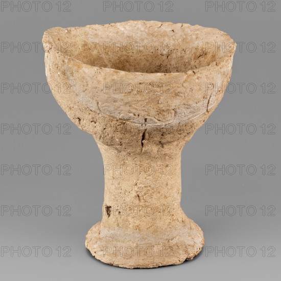 Beaker with engraved decoration, unknown northern Mesopotamian workshop. Hand-made beaker on a thick, cylindrical stem with a poorly defined foot. The bowl is a low form with an irregularly defined rim, glued to the foot. Three schematically engraved human figures on the bowl: the torso with head and legs shown by a vertical line ending in a dot at the top and a short downward-opening arc at the bottom; arms extended upward marked by an engraved horizontal line curved at both ends, intersecting the torso slightly below midpoint. Anthropomorphic depictions; engraved decorations; Near Eastern ceramics; Eastern Gaulish Scenery. 1st half of the 3rd millennium BC; Early Jazira III period Early Bronze/Early Dynastic III; ca. 2600–ca. 2350 BC. Findspot: Tell Abu Hafur northeastern Syria, Khabur basin, Northern Mesopotamia Khabur basin. Vessel, weight 148 g., A rough, bowl-shaped ceramic object with a wide, shallow upper cavity atop a narrower cylindrical stem and a flared base, showing an une...