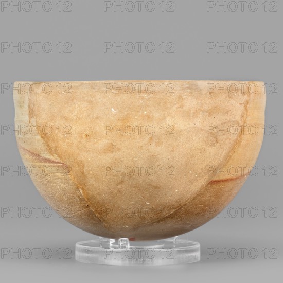 Bowl, unknown Northern Mesopotamian workshop. A hemispherical bowl topped with a simple rim, diagonally cut inward. On the outside, below the rim, a wide, shallow groove runs. The diagonal pink line and the darker surface of the lower part of the bowl are a technological trace created during firing. 1st half of the 3rd millennium BC; Early Jazira III period ca. 2600–ca. 2350 BC. Tell Abu Hafur North-Eastern Syria, Northern Mesopotamia Khabur basin. Ceramic vessel; height 6.5 cm, width 10 cm, weight 136 g., A rounded, shallow bowl displayed on a clear circular stand, with a smooth, matte surface in shades of tan and beige with a few subtle reddish streaks and small pale speckles; the background is a flat gray.