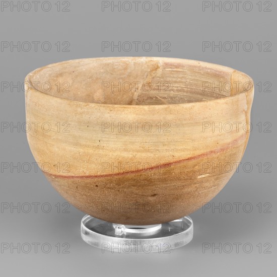A hemispherical bowl topped with a simple rim, diagonally cut inward. On the outside, below the rim, a wide, shallow groove runs. The diagonal pink line and the darker surface of the lower part of the bowl are a technological trace created during firing. Unknown Northern Mesopotamian workshop. 1st half of the 3rd millennium BC; Early Jazira III period ca. 2600–ca. 2350 BC. Tell Abu Hafur north-eastern Syria, Northern Mesopotamia Khabur basin. Vessel; height 6.5 cm, width 10 cm, weight 136 g., A rounded bowl resting on a clear circular stand, made of a stone-like material with a smooth matte surface showing layered bands and subtle striations; the bowl displays a range of beige and tan tones with areas of light brown and a thin reddish-brown streak around its midsection, and the interior shows similar beige and tan shading with faint concentric markings.