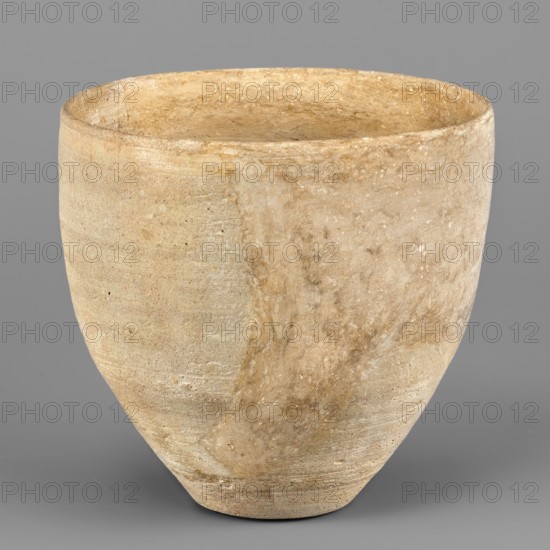 Tall bowl, unknown Northern Mesopotamian workshop. Tall bowl with a small, flat bottom. The walls of the vessel, tilted at the bottom, become vertical at 1/3 of their height, terminating in a straight rim separated from the outside by a shallow, engraved line. 1st half of the 3rd millennium BC; Early Jazira III period Early Dynastic III; ca. 2600–ca. 2350 BC. Tell Abu Hafur North-Eastern Syria, Northern Mesopotamia Khabur basin. Vessel; height 11 cm, width 11.6 cm, weight 250 g., A roughly rounded vessel with a wide open top and a tapered base sits against a plain background; the surface shows a textured, slightly rough finish with visible striations and patches of wear; colors visible include beige, tan, light brown, and hints of off-white and gray; the rim appears slightly uneven and the interior shows similar coloration and texture to the exterior; overall the shape is symmetrical with a subtle vertical variation in tone around the body.