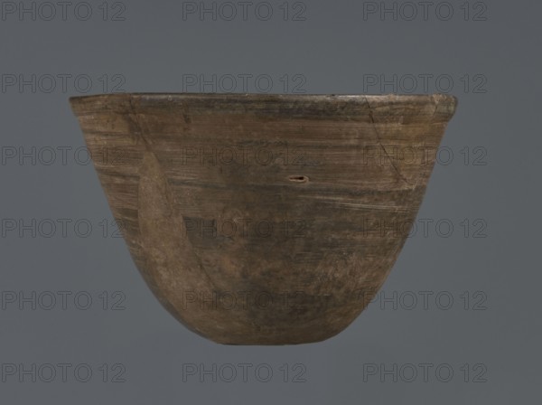 Bowl, unknown northern Mesopotamian workshop; metallic ware, viceless vessel, bowl; Early Jazira III period ca. 2600–ca. 2350 BC / Early Dynastic III period ca. 2600–ca. 2350 BC; metallic ware type bowl with a small, flat bottom and straight, tilted walls ending with a straight, slightly thickened, outwardly curled rim; ca. 2600–ca. 2350 BC; Tell Abu Hafur northeastern Syria, Northern Mesopotamia Khabur basin, Al-Jazira Syria/Iraq/Mesopotamia; vessel; height 7.5 cm, width 11.85 cm, bottom diameter 3.2 cm., A brown, rounded ceramic bowl with a flared rim is centered against a solid gray background; the bowl shows a worn, uneven surface with visible horizontal striations, patches of darker and lighter brown, and an area of smoother, lighter brown on one side, along with small chips, a narrow hole, and faint scratches.