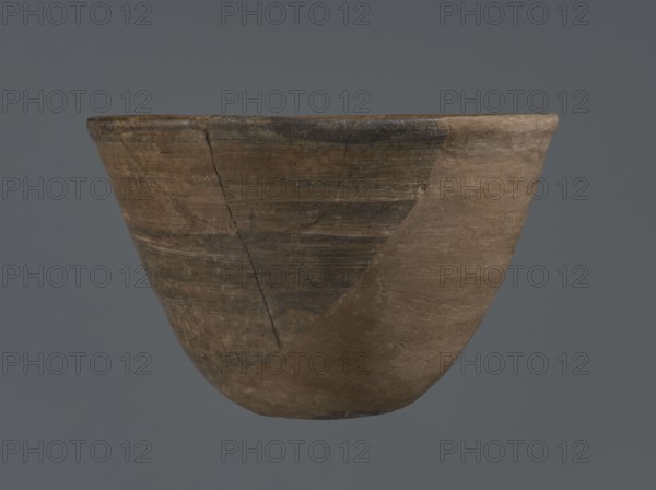 Bowl, unknown northern Mesopotamian workshop, metallic ware, viceless vessel, bowl, Early Jazira III period ca. 2600–ca. 2350 BC / Early Dynastic III period ca. 2600–ca. 2350 BC. Metallic ware type bowl with a small, flat bottom and straight, tilted walls ending with a straight, slightly thickened, outwardly curled rim. Ca. 2600–ca. 2350 BC. Tell Abu Hafur northeastern Syria, Northern Mesopotamia Khabur basin. Vessel; height 7.5 cm, width 11.85 cm, bottom diameter 3.2 cm., A worn, rounded ceramic bowl floats against a gray background, showing a surface of earthy browns and darker brown patches with visible cracks and repaired joins; the rim is slightly irregular and darker along one section, and the bowl’s texture appears rough and matte with subtle horizontal striations and areas of lighter beige-brown.