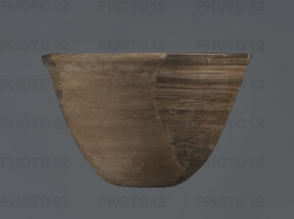 Bowl, unknown northern Mesopotamian workshop; metallic ware; viceless vessel; bowl; Early Jazira III period ca. 2600–ca. 2350 BC; Early Dynastic III; Al-Jazira Syria/Iraq, Mesopotamia; metallic ware type bowl with a small flat bottom and straight, tilted walls ending in a straight, slightly thickened outwardly curled rim; ca. 2600–ca. 2350 BC; Tell Abu Hafur northeastern Syria, Northern Mesopotamia Khabur basin; vessel; height 7.5 cm, width 11.85 cm, bottom diameter 3.2 cm., A ceramic bowl shown against a gray background, with a rounded body and flared rim, displaying an overall earthy brown color with areas of darker brown and muted tan; the right side of the bowl shows horizontal striations and a slightly darker, weathered texture while the left side appears smoother and more uniformly brown; the rim has a thin darker band and the surface shows subtle irregularities, scratches, and patches of lighter and darker brown tones.