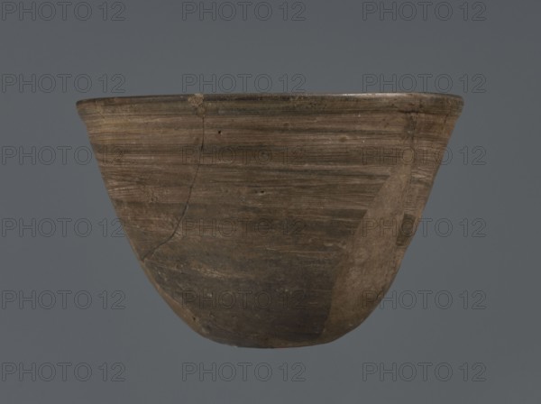 Bowl, unknown northern Mesopotamian workshop; metallic ware; viceless vessel; bowl; Early Jazira III period ca. 2600–ca. 2350 BC / Early Dynastic III period ca. 2600–ca. 2350 BC; Al-Jazira Syria; Iraq; Mesopotamia; metallic ware type bowl with a small, flat bottom and straight, tilted walls ending with a straight, slightly thickened, outwardly curled rim; ca. 2600–ca. 2350 BC; Tell Abu Hafur northeastern Syria, Northern Mesopotamia Khabur basin; vessel; height 7.5 cm, width 11.85 cm, bottom diameter 3.2 cm., A worn bowl with a rounded base and flared rim sits against a plain gray background; the bowl shows a weathered surface with visible horizontal striations and patches of smoother repair, and its colors include shades of brown, tan, and darker brown areas with subtle olive and grayish tones.