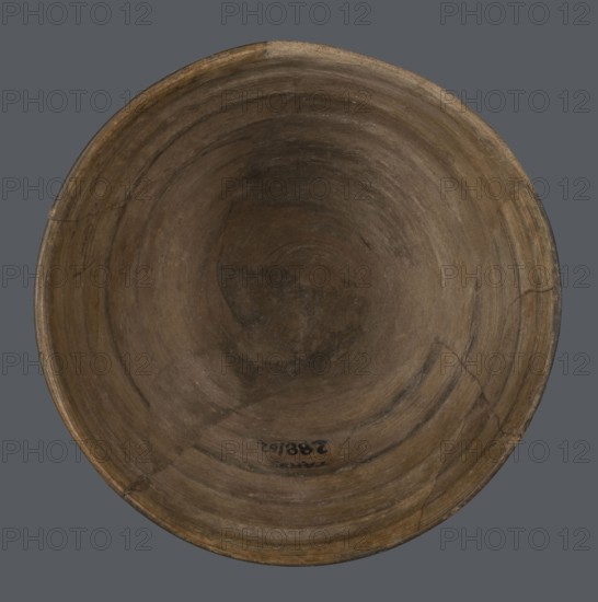Bowl, unknown northern Mesopotamian workshop; metallic ware; viceless vessel; bowl; Early Jazira III period ca. 2600–ca. 2350 BC; Early Dynastic III period; metallic ware type bowl with a small flat bottom and straight, tilted walls ending with a straight, slightly thickened, outwardly curled rim; ca. 2600–ca. 2350 BC; Tell Abu Hafur northeastern Syria, Northern Mesopotamia Khabur basin; vessel; height 7.5 cm; width 11.85 cm; bottom diameter 3.2 cm., A round shallow bowl viewed from above with a worn surface showing concentric circular ridges and cracks, displaying shades of brown and tan with darker brown patches and subtle blackish areas, set against a flat gray background.