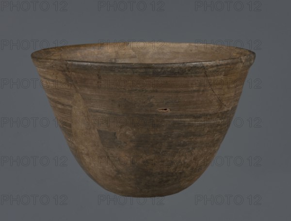Bowl, unknown northern Mesopotamian workshop; metallic ware; viceless vessel; bowl; Early Jazira III period ca. 2600–ca. 2350 BC / Early Dynastic III period; Al-Jazira Syria/Iraq/Mesopotamia; metallic ware type bowl with a small, flat bottom and straight, tilted walls ending with a straight, slightly thickened, outwardly curled rim; ca. 2600–ca. 2350 BC; Tell Abu Hafur northeastern Syria, Northern Mesopotamia Khabur basin; vessel; height 7.5 cm; width 11.85 cm; bottom diameter 3.2 cm., A worn, rounded ceramic bowl sits against a plain gray background, showing a surface of earthy browns and muted tans with darker brown streaks and patches; the rim has a slightly darker brown band and the bowl displays visible repair marks and hairline cracks, along with uneven texture and areas of lighter beige where the material appears smoother.