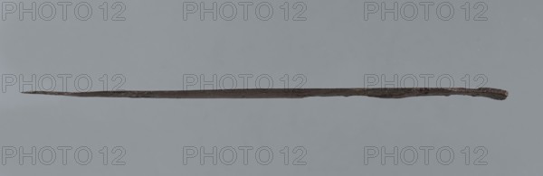 Dagger blade, unknown Mitannian workshop; weapons, daggers; Mitannian period ca. 1550–ca. 1250 BC. The blade of a short dagger with a centric point. At the upper end it terminates in a short, rectangular base blunted on both sides, behind which the double-edged blade tapers evenly towards the tip. Hilt not preserved. c. 1550–c. 1250 BC; Tell Abu Hafur East northeastern Syria, Northern Mesopotamia Khabur basin. Height 15.6 cm; blade width 2.5 cm; depth 0.2 cm; base length 1.7 cm; base width 1.7 cm., A long, thin, dark brown metallic object lies horizontally across a muted gray background, tapering to a pointed end on one side and appearing slightly thicker and irregular on the opposite side; the surface shows rough, textured areas and subtle variations in brown and darker gray tones.